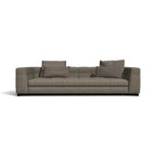 A modern three-seater sofa upholstered in textured taupe-gray fabric, featuring four matching throw pillows and a low black base, shown against a plain white background.