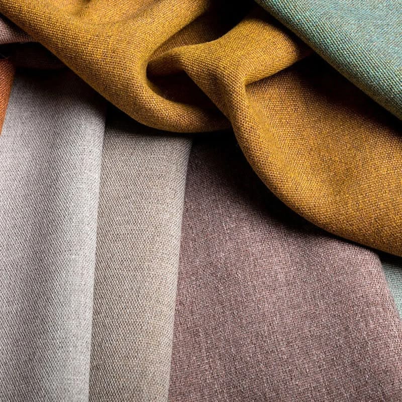 Close-up of draped woven fabric in multiple soft colors, including mustard yellow, dusty rose, pale gray, and muted sage green, with visible textile texture.